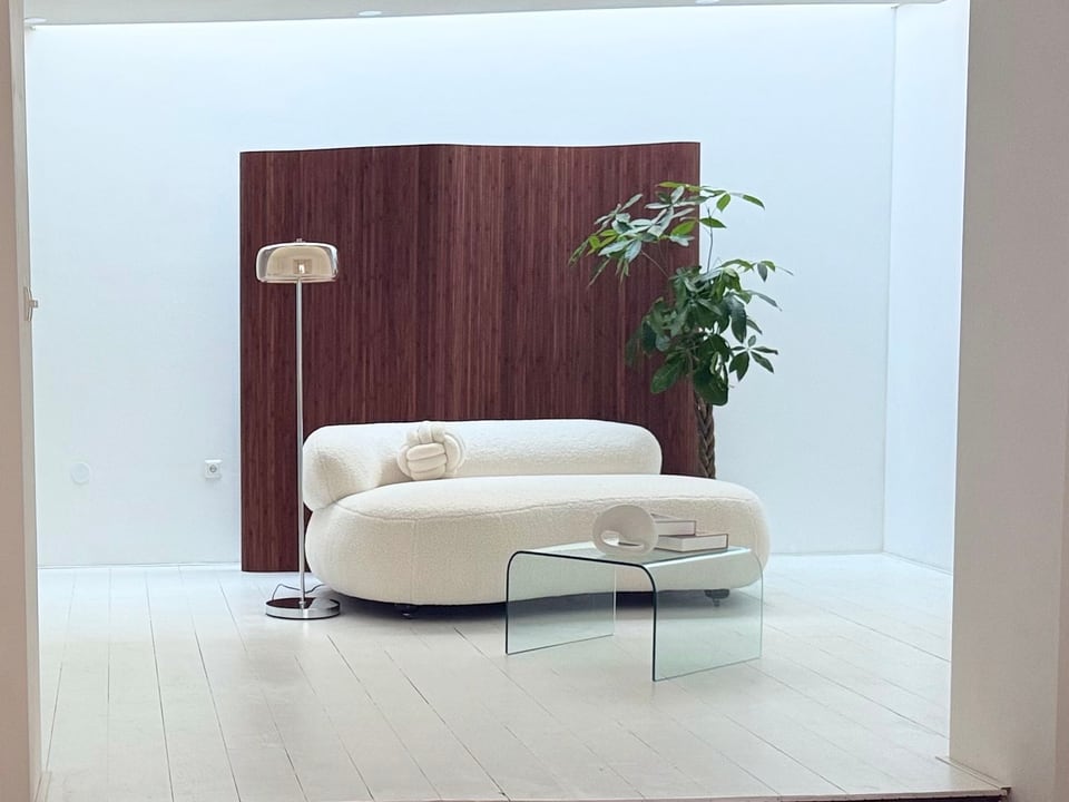 De studio gezien vanaf de trap: een lichte nis met een golvend houten kamerscherm, een witte bank, een glazen salontafel, een vloerlamp en een plant